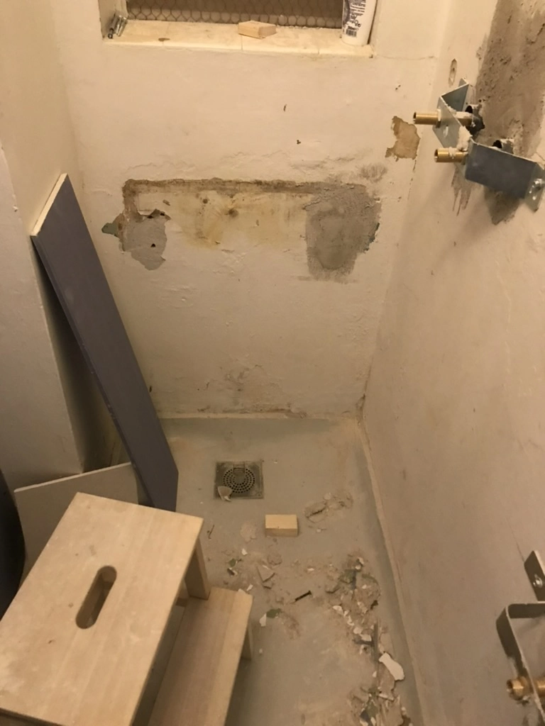 Badeværelse under renovering med bar væg, synlige vandrør, fjernede fliser og ujævnt gulv. Renovering af badeværelse