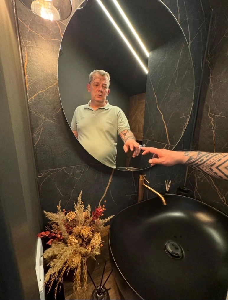 Mand i spejl over sort vask med guldfarvet vandhane og mørke marmorerede fliser – renovering af badeværelse