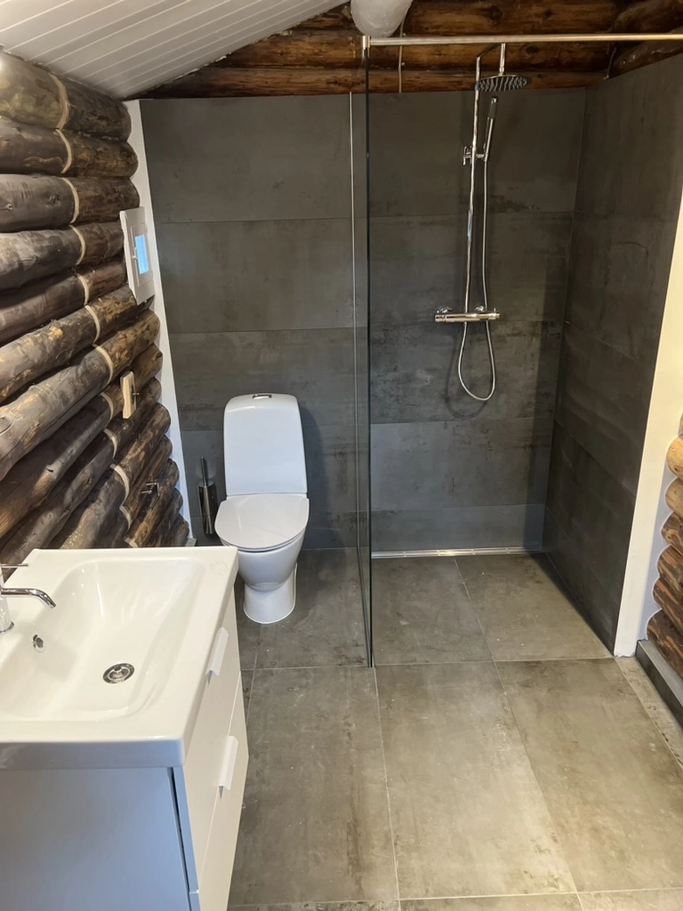 Renoveret vådrum med store grå fliser på gulv og væg, glas bruseniche, hvidt toilet og hvidt skabsbassin - Renovering af badeværelse