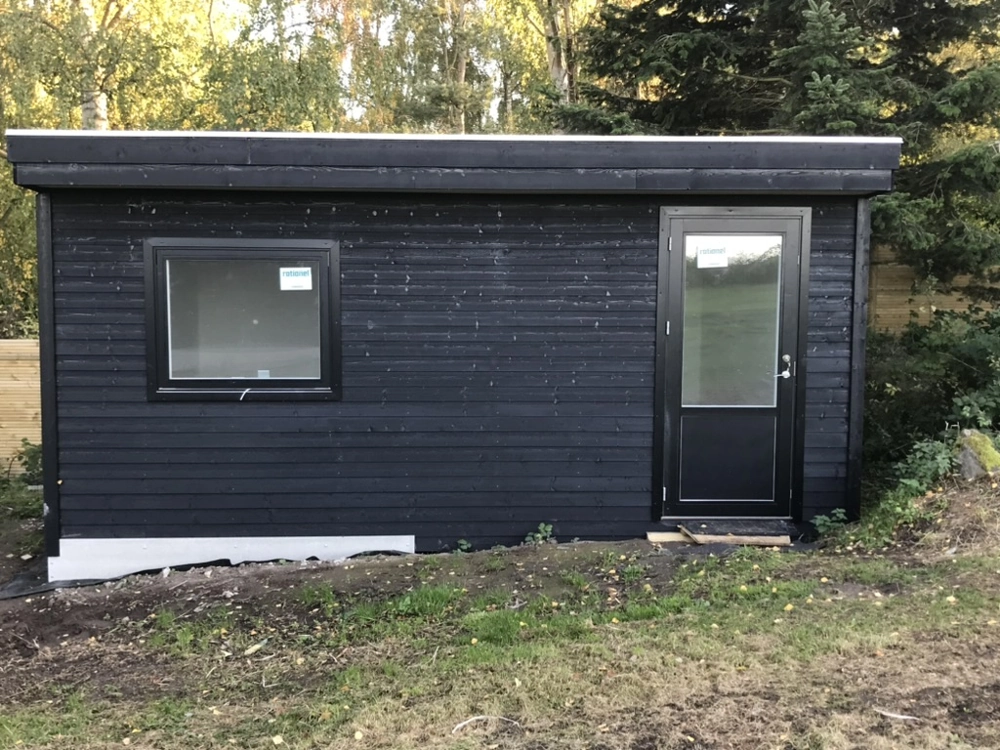 Sort træhus med fladt tag, enkelt vindue og dør, placeret på græs og omgivet af træer