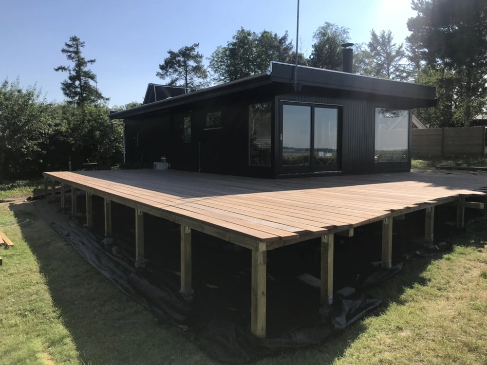Ny træterrasse monteret på stolper foran moderne sort facadehus med bredt plankegulv