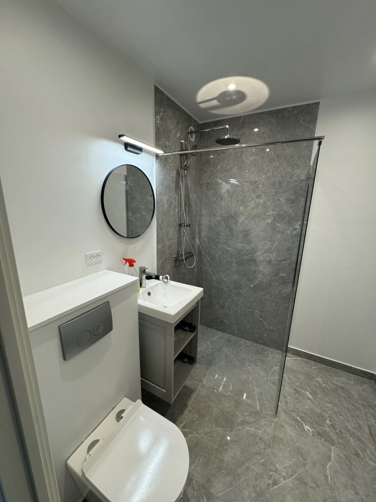Renovering af badeværelse med grå fliser, glasbrusevæg, væghængt toilet, vaskeskab og rundt spejl